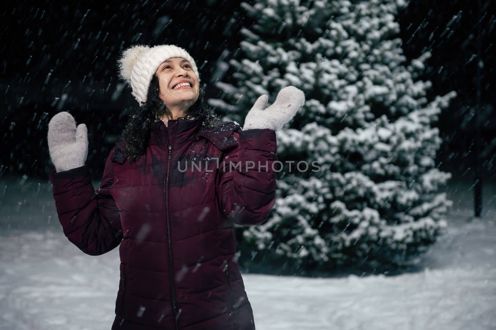 Delightful happy beautiful woman in white wool mittens and hat, having fun, catching snowflakes, standing outdoors on a snow covered nature on a snowy cold winter evening. Christmas holidays concept