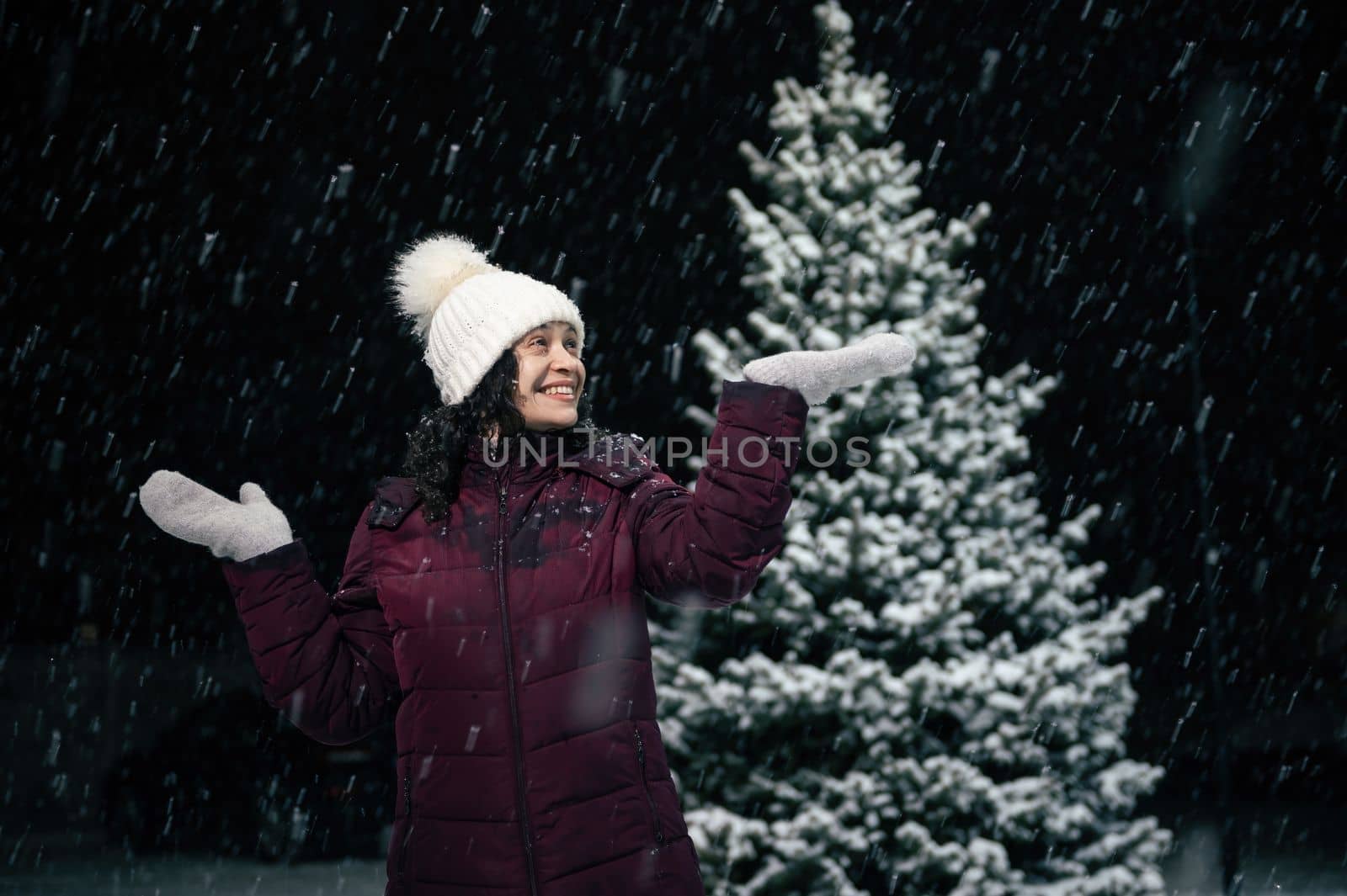 Charming African American woman in white wool hat and mittens, experiencing happiness, catching snowflakes falling on her hands, on a snowy winter evening, standing against a snow covered pine tree