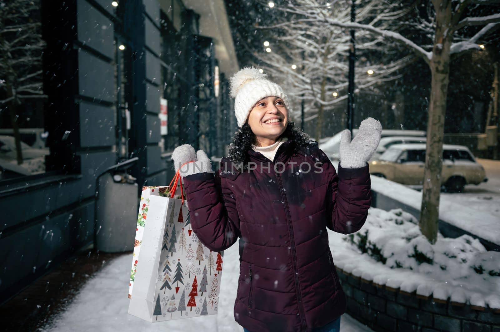 Delightful young multi-ethnic woman in warm clothes, catches snowflakes while walking down the snow covered city street with shopping bags, enjoying happy winter holidays on a snowy cold night