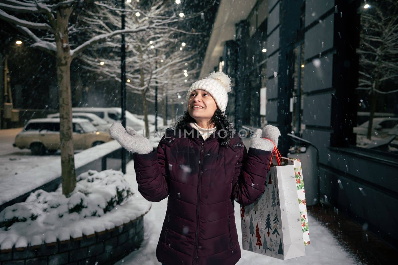 Delightful young multi-ethnic woman in warm clothes, holding shopping bags, catching snowflakes while walking down the snow covered city street, illuminated by festive garlands on a snowy cold night