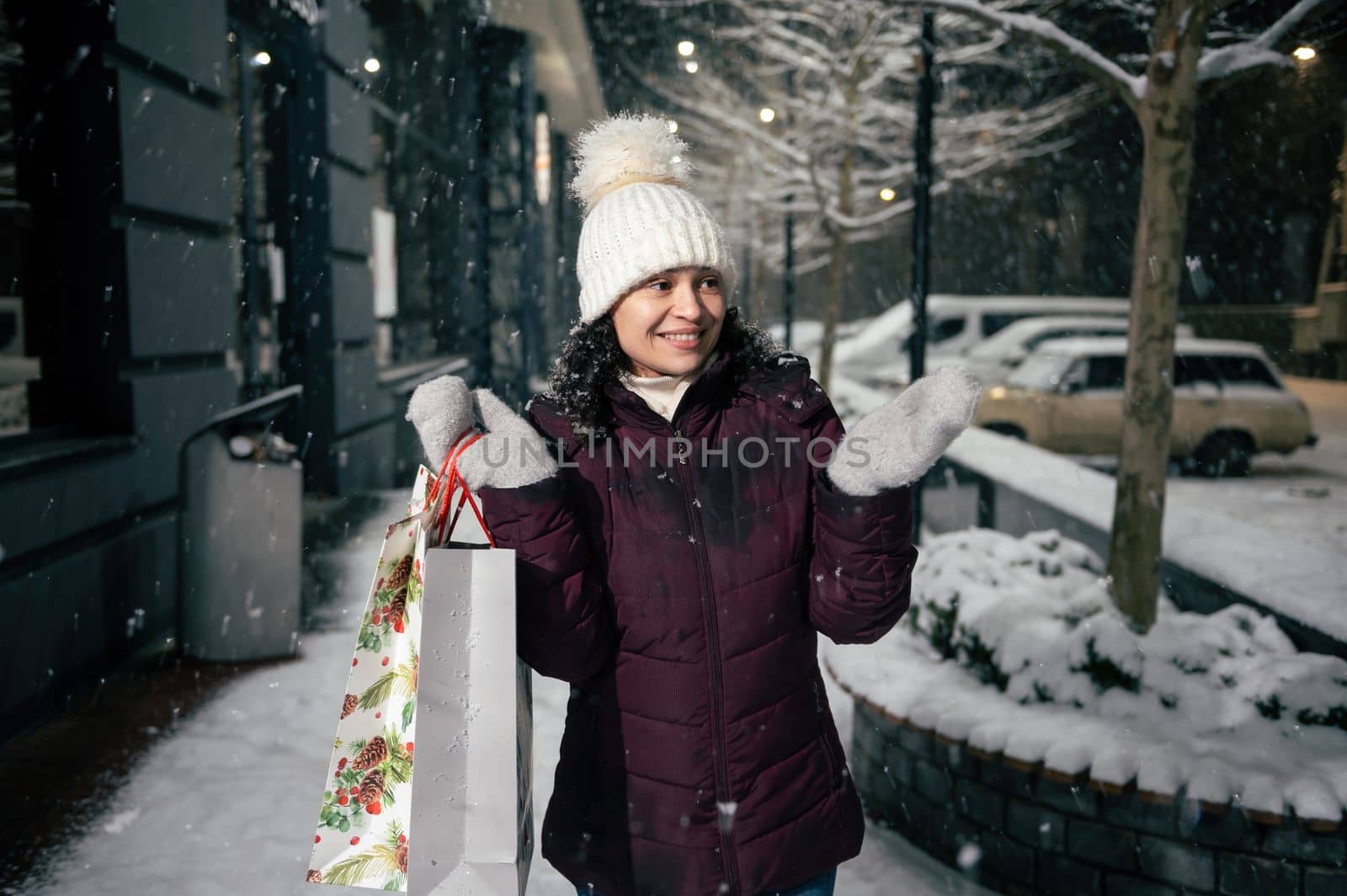 Attractive multi-ethnic young woman in warm clothes, with shopping bags, catches snowflakes falling on her hands while walking along a snowy street illuminated by holiday garlands. Merry Christmas