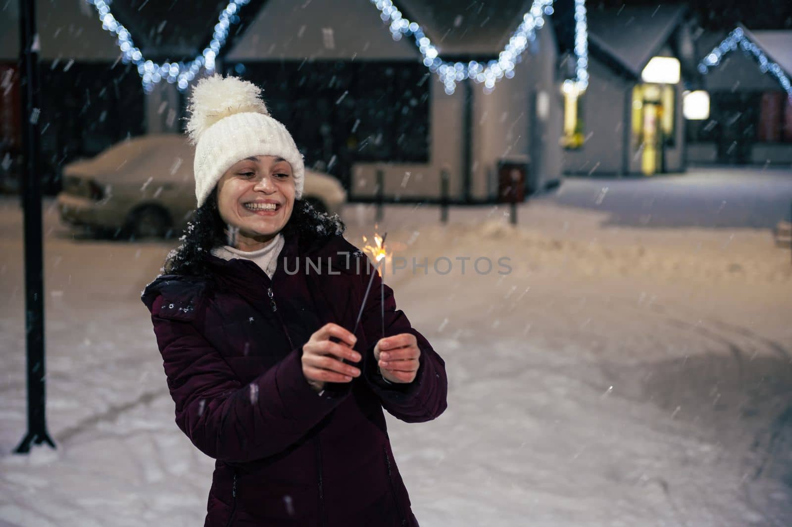 Charming young cheerful woman in warm winter clothes, lights sparklers on a snow-covered street lit by New Year's garlands. Snow falls on a beautiful winter evening. Merry Christmas concept