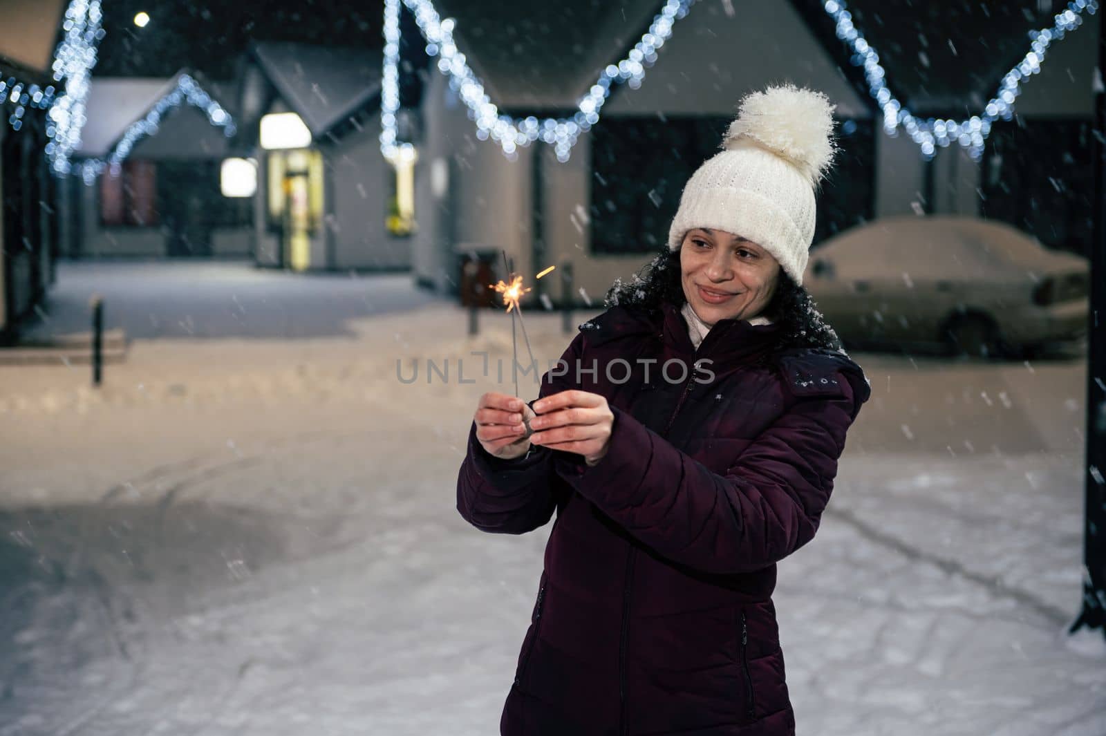 Pretty multi-ethnic woman lighting sparklers, Bengal lights while walking on a city street, illuminated by garlands on a snowy evening. Enjoying winter and snow covered urban environment