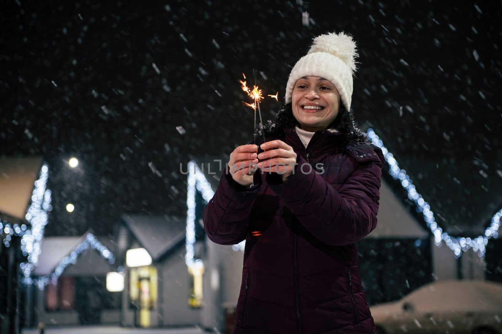 Delightful woman lighting sparklers, Bengal lights on a city street, illuminated by garlands on a snowy evening. Merry Christmas concept. Winter holidays.