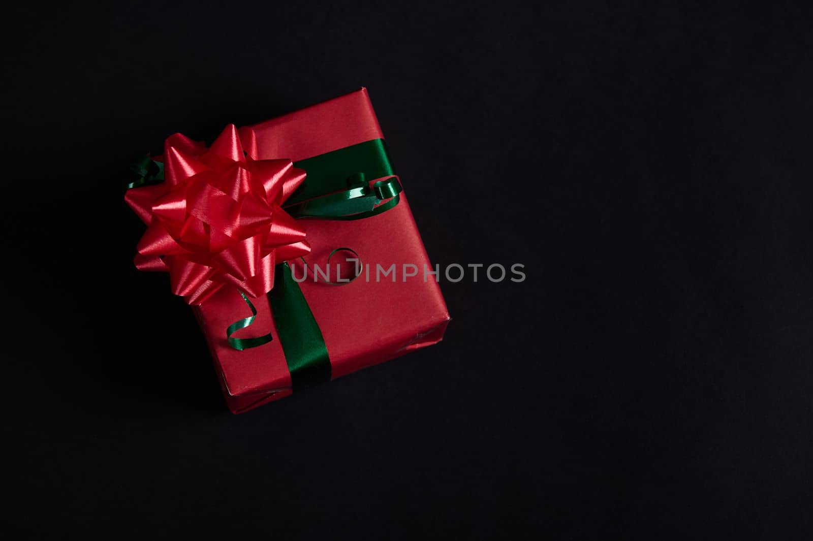 Still life with a Christmas present in a gift box wrapped in red wrapping paper, with green ribbon and red tied bow on black background. Flat lay. December 25, Happy New Year, Christmastime