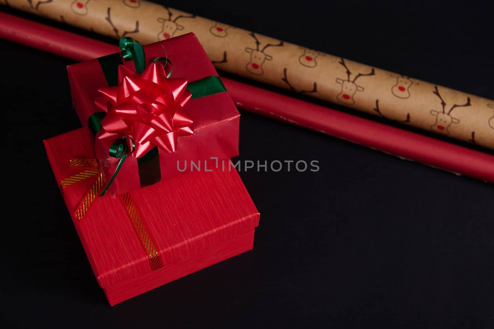 Still life with gift boxes, ornate with green ribbon and red tied bow, surrounded by wrapping paper on a black background. Copy space. Happy New Year. Merry Christmas. Winter holidays. Christmastime