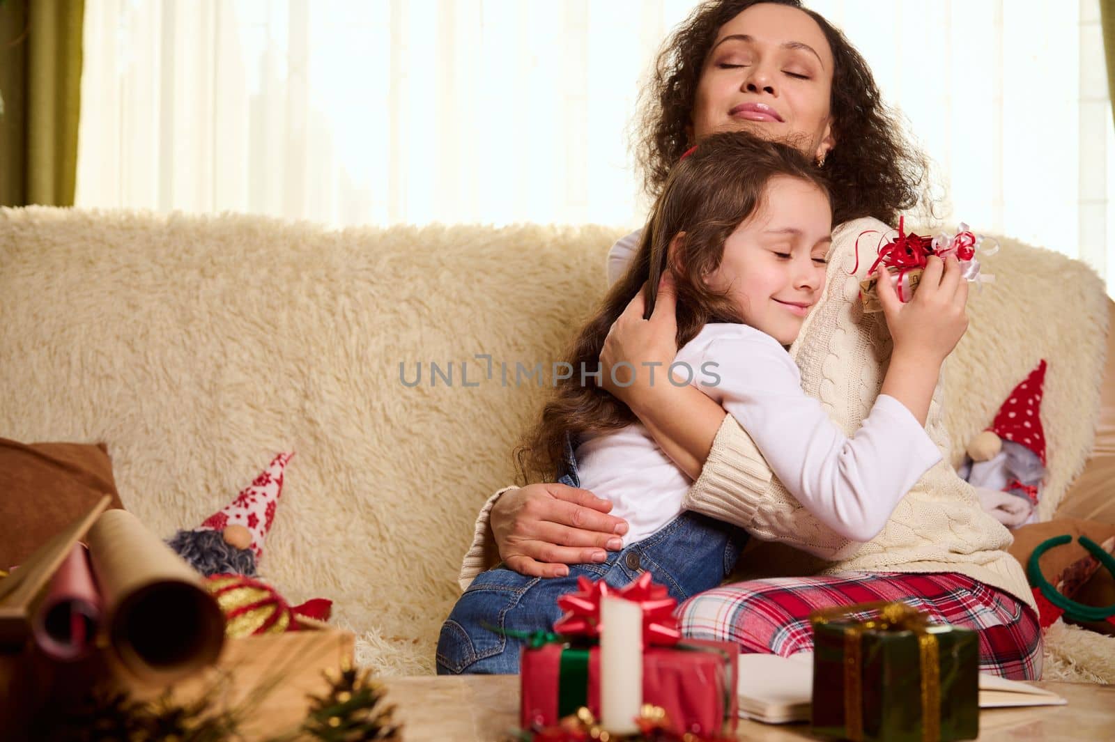 Charming little girl, beloved daughter gently leans on the shoulders of her mother hugging her, receiving a cute Christmas present. Beautiful gifts on the table in foreground. Happy carefree childhood