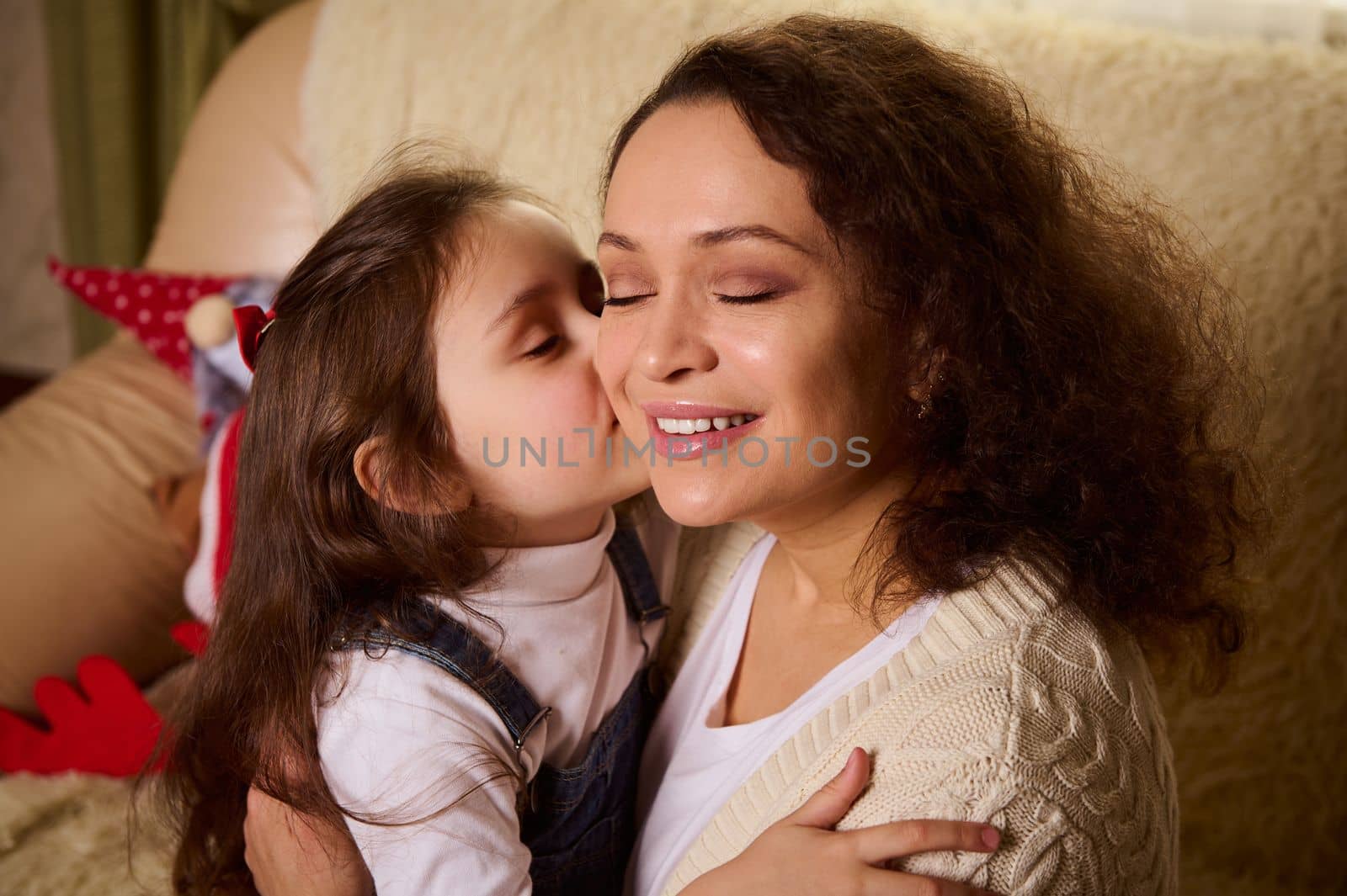 Multi-ethnic beautiful delightful young woman, a loving mother, experiences happiness when her adorable daughter hugs and kisses her on cheek. Happy family relationships. Mother's Day. Children's Day