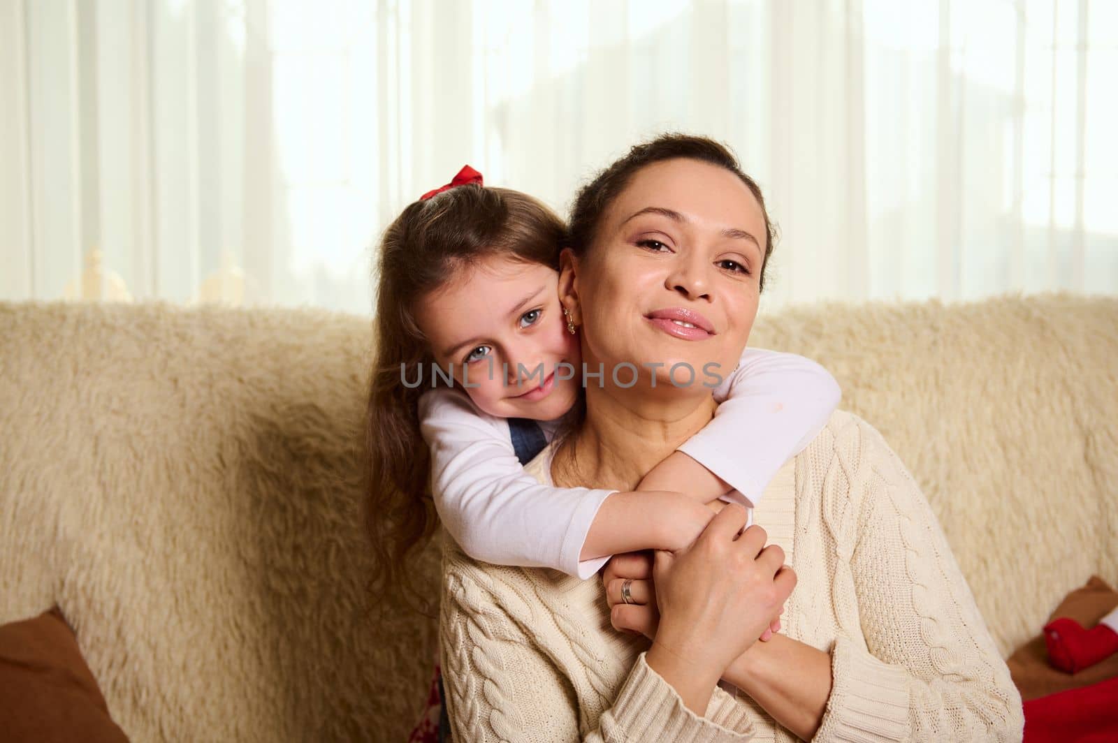 Lovely little girl gently hugging her mother from the back, sitting together on sofa at home and cutely smiling looking at camera. Happy Childhood and Motherhood. Love. Care. Family relationships
