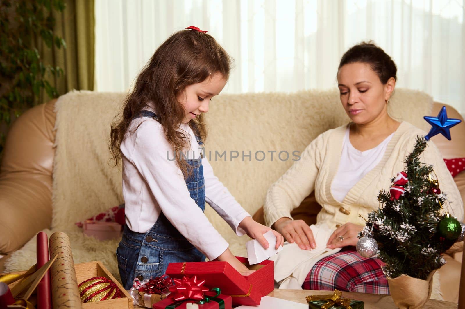 Beautiful multi-ethnic woman, loving mother and her adorable little daughter packing presents for Christmas, doing New Year preparations. Magical atmosphere of winter holidays. Xmas. December 25th