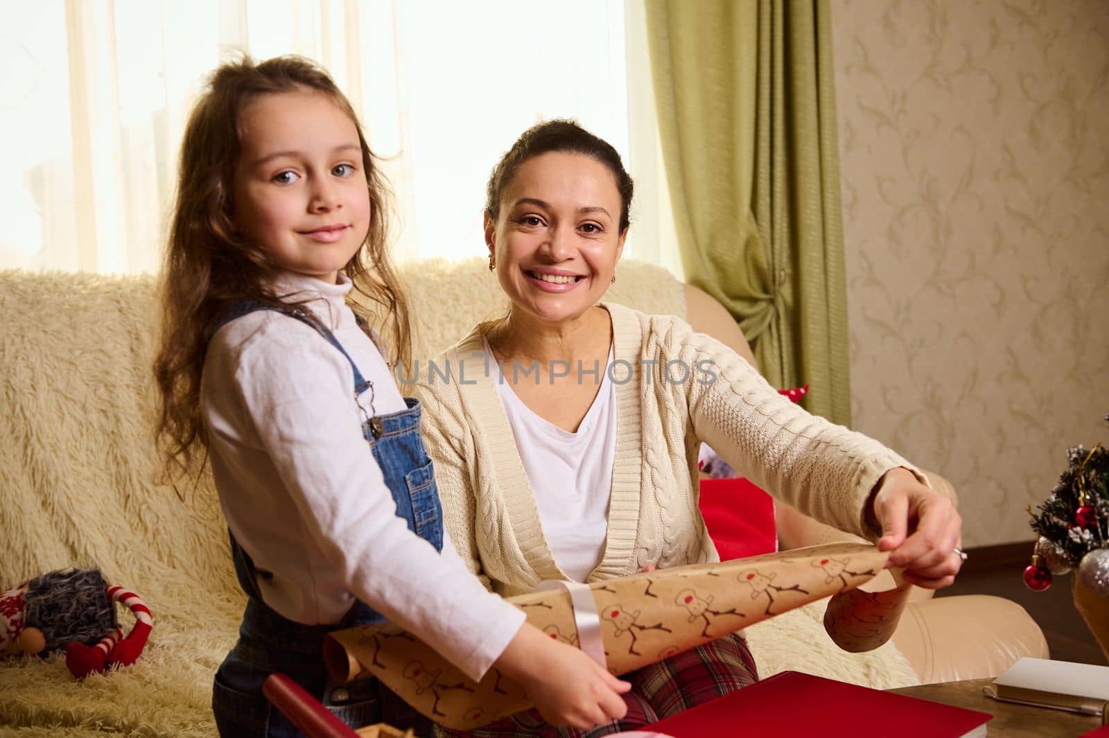 Pretty woman, loving mother and lovely daughter smile sweetly looking at the camera while wrapping gifts for Christmas, New Year or any other occasion. Looking forward to the upcoming winter holidays