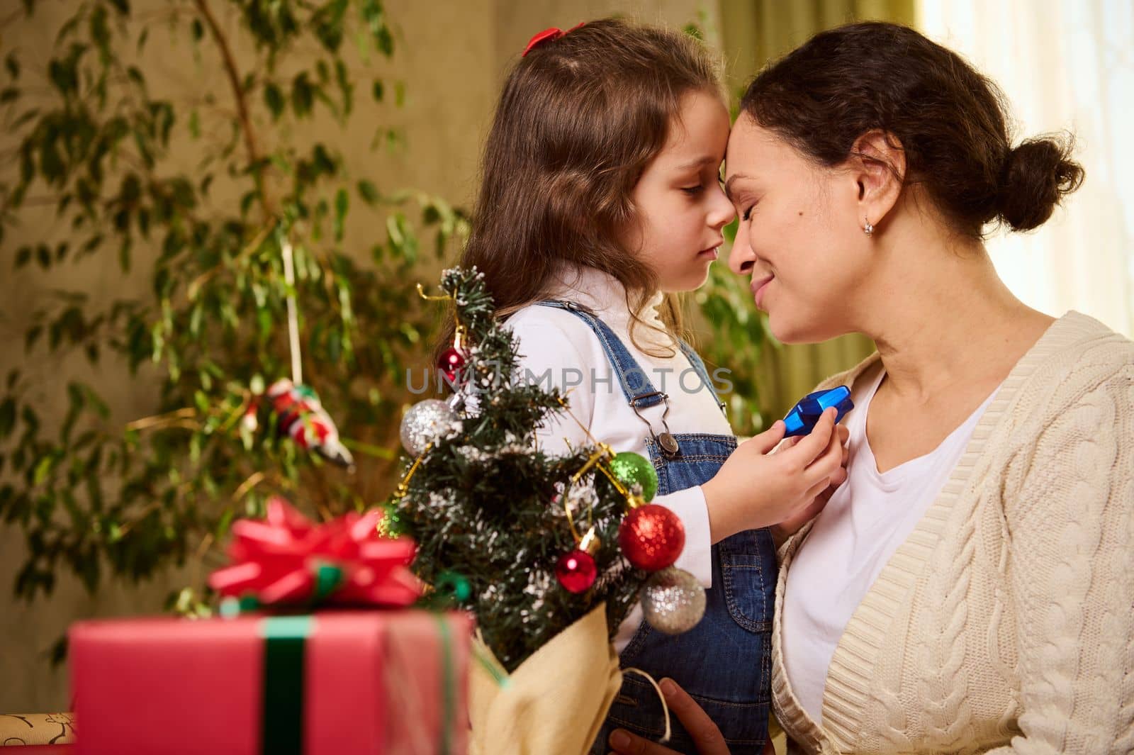 Happy woman, loving affectionate mother and her beloved little daughter enjoying togetherness, hugging each other while decorating Christmas tree at home. Blurred beautiful presents on the foreground