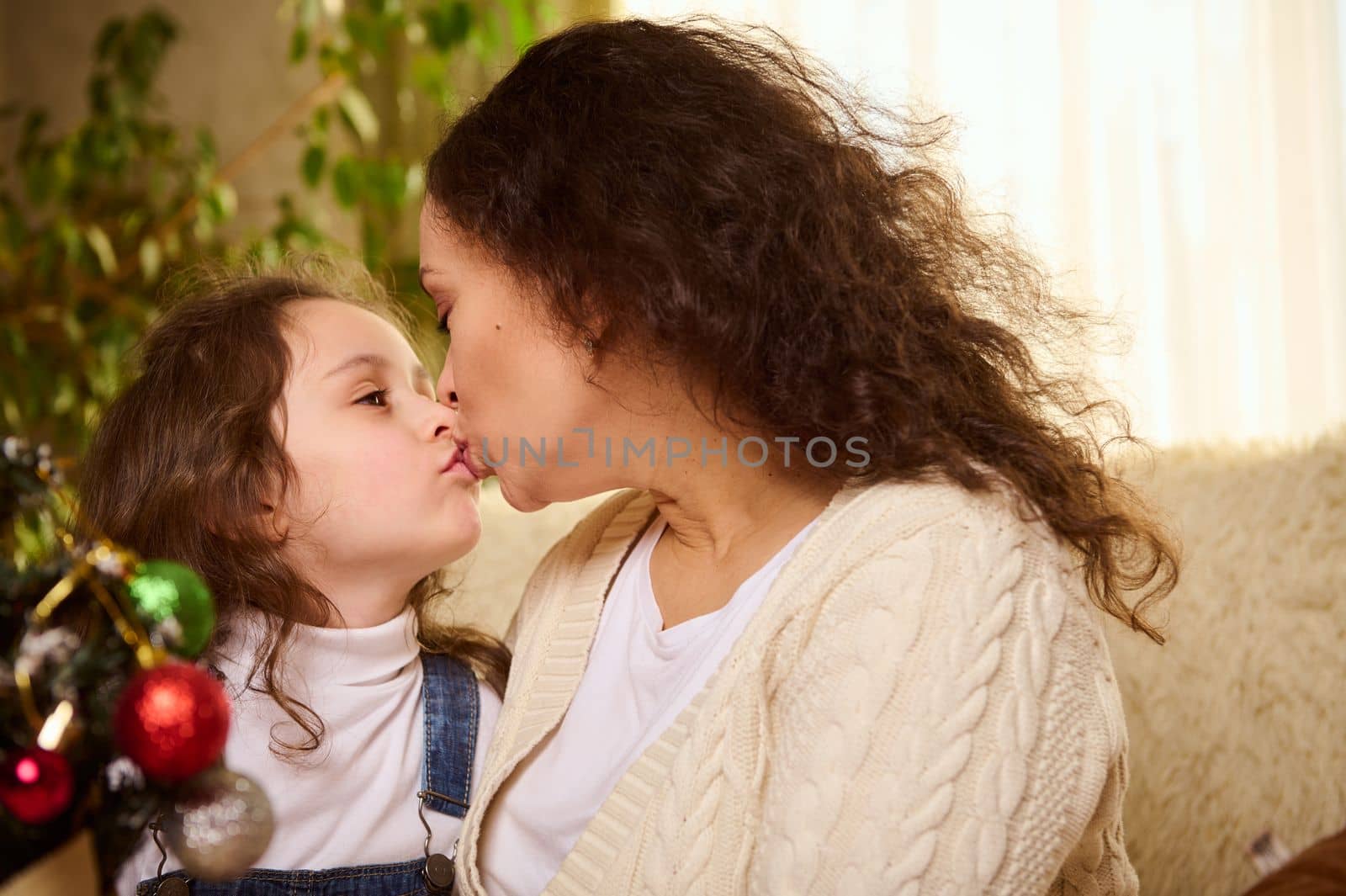 Adorable Caucasian child, a little girl, lovely daughter kissing her affectionate loving caring mother while decorate together a Christmas tree. Happy New Year. Magic atmosphere of winter holidays