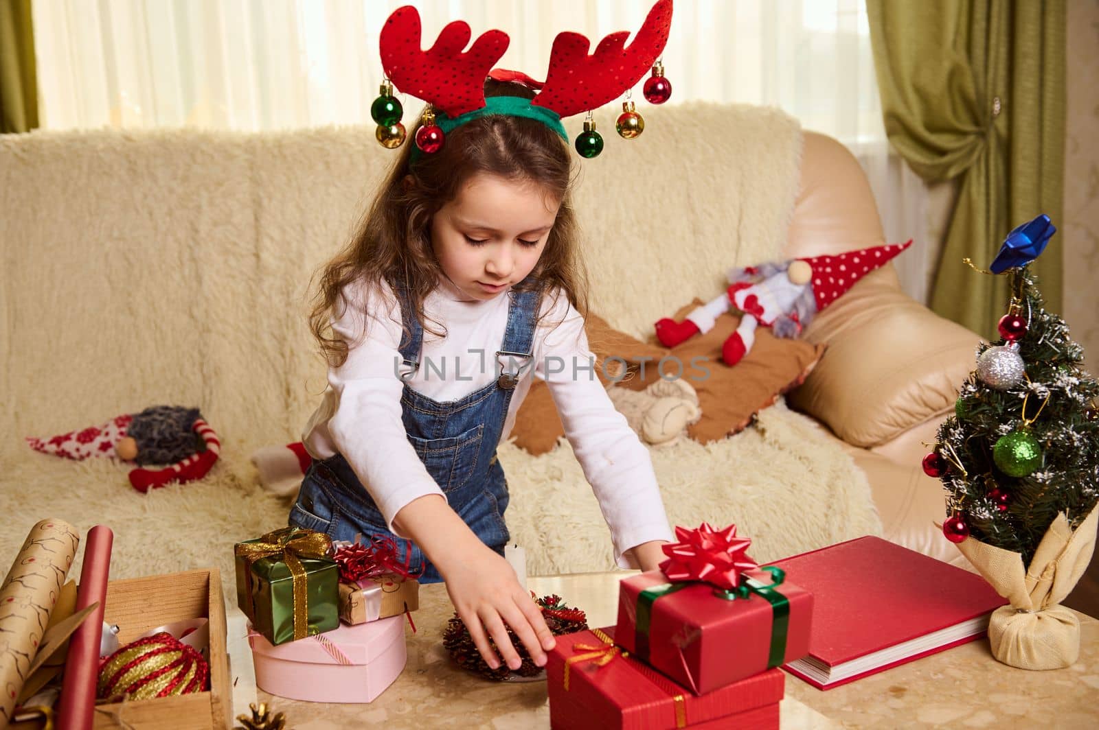 Adorable Caucasian little girl wearing deer antler hoop and denim overalls, standing at a table with Christmas gifts, wrapping materials for packing presents. Xmas. Happy New Year and winter holidays