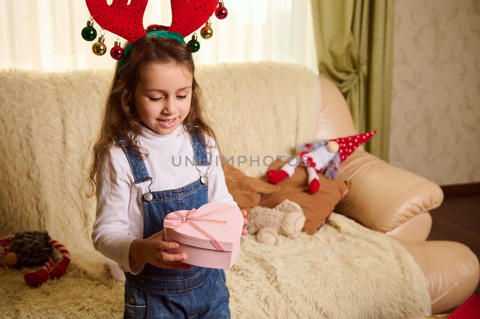 Cute little child, baby girl holds a pink heart shaped gift box in her hands. Happy New Year. Merry Christmas. Boxing Day. Beautiful presents for upcoming festive events. Atmosphere of winter holidays