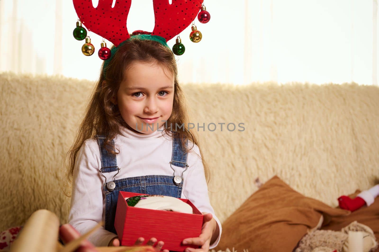 Cute baby wearing deer antler hoop, smiles looking at camera while unpacking a beautiful red gift box. Boxing Day. Happy magic atmosphere of winter holidays. New Year. Merry Christmas. Christmastime