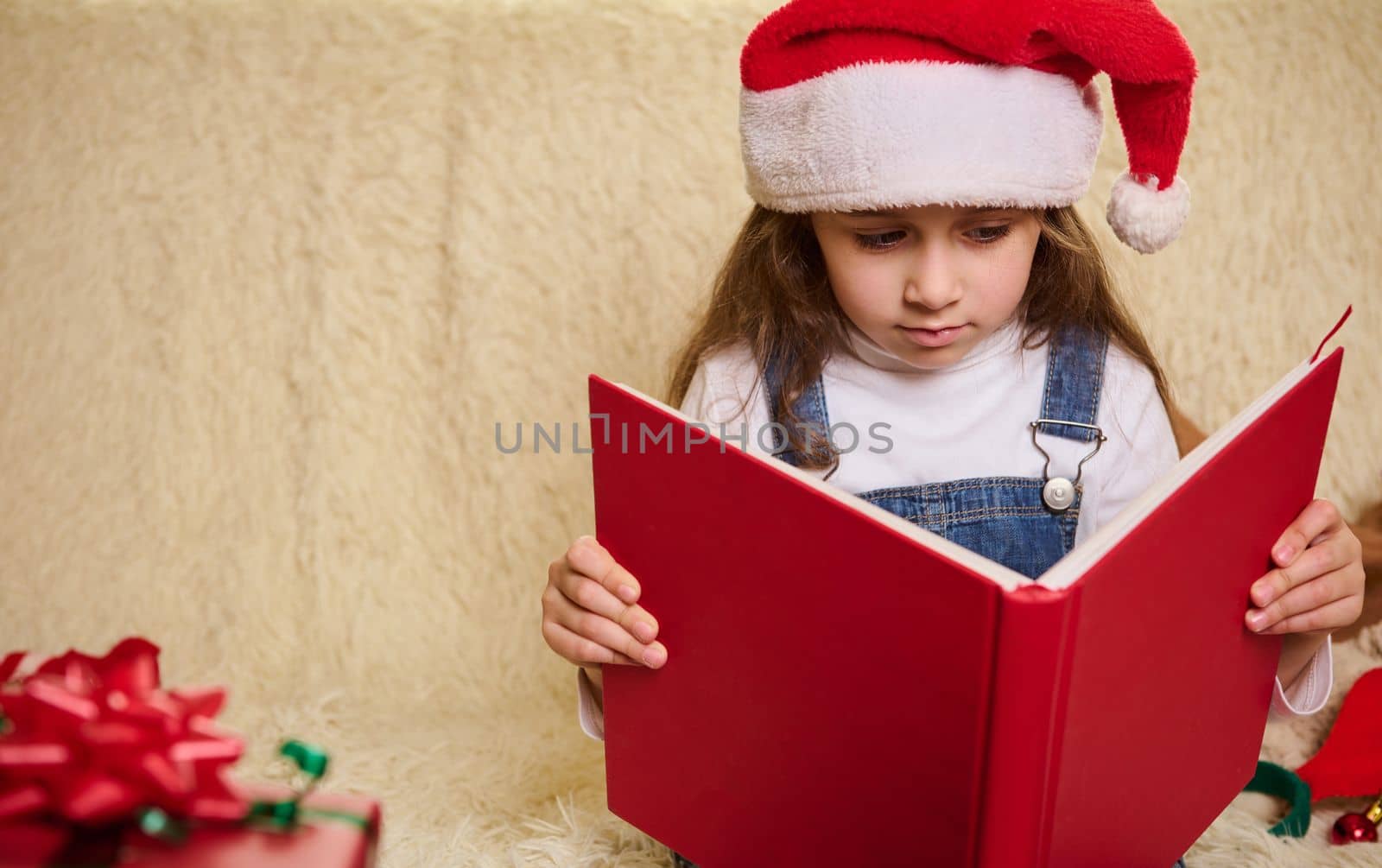 Portrait of a cute little child girl in Santa's hat, with a hardcover red book, reading fairy tales at cozy home interior with Christmas decors. Copy advertising space for promotional text. New Years