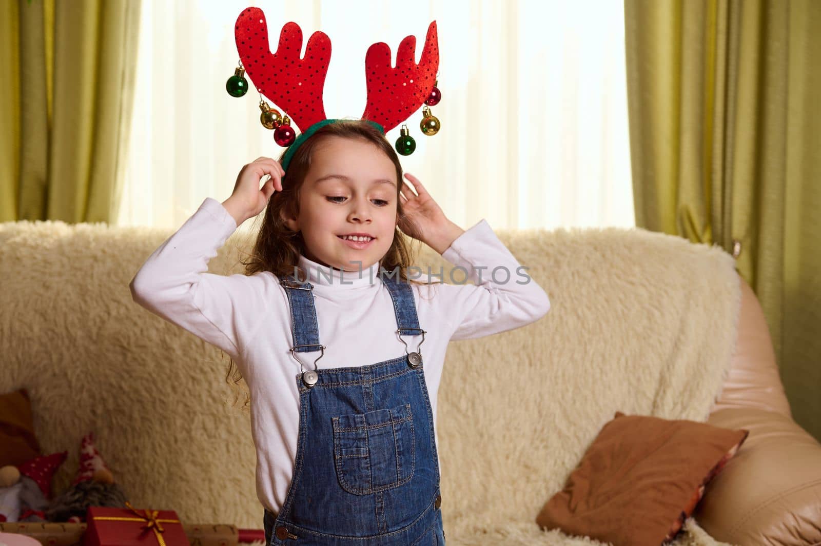 Adorable child, a Caucasian little girl in denim overalls and white pull, puts on antler deer hoop, enjoys Christmas atmosphere at home. Happy winter holidays. Magic mood. December 25. Happy childhood