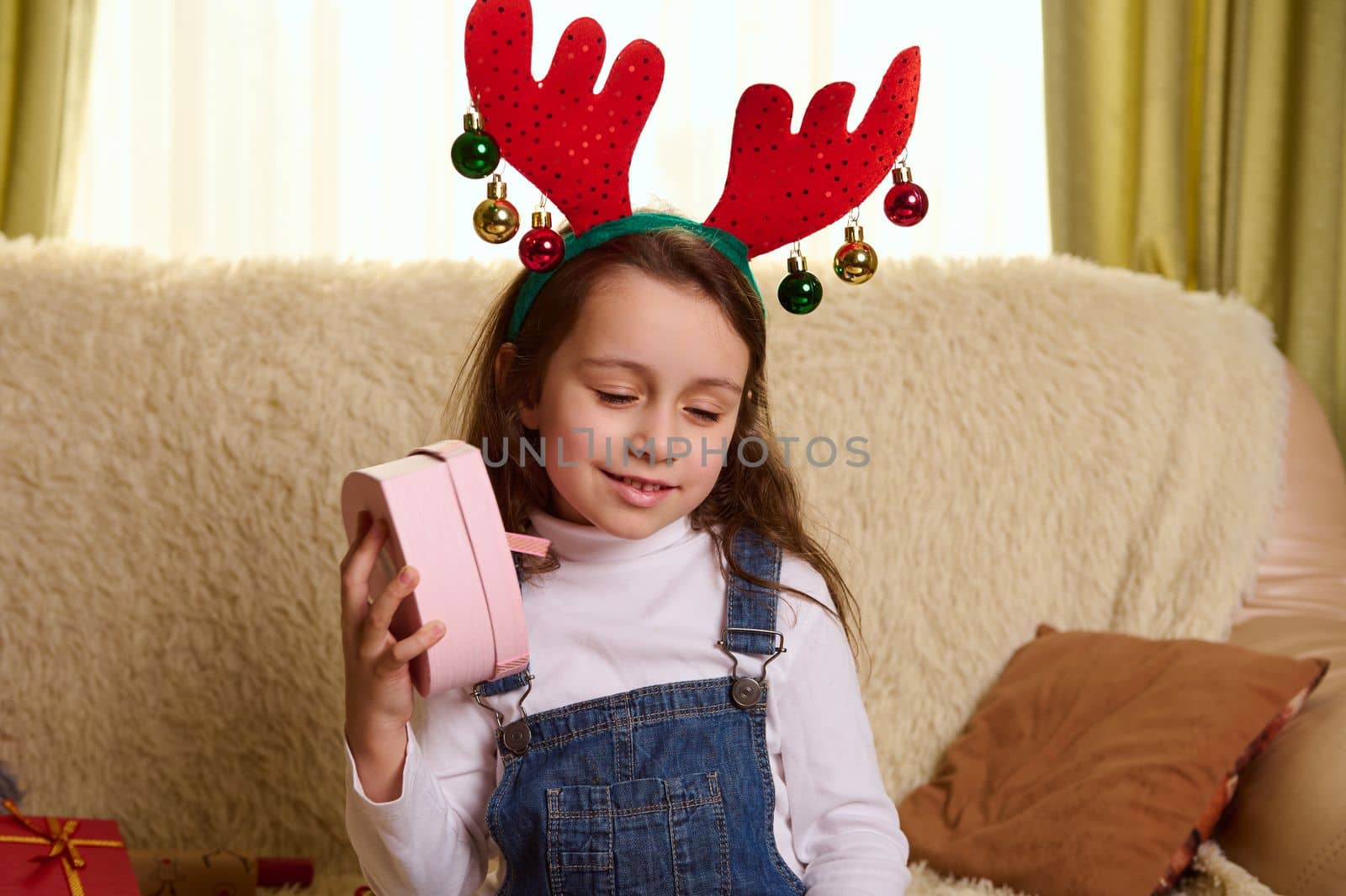 Charming cutie, adorable European little girl in deer antlers hoop, shakes a pink heart shaped gift box, trying to guess its contents. Happy surprises and presents for Christmas or New Year