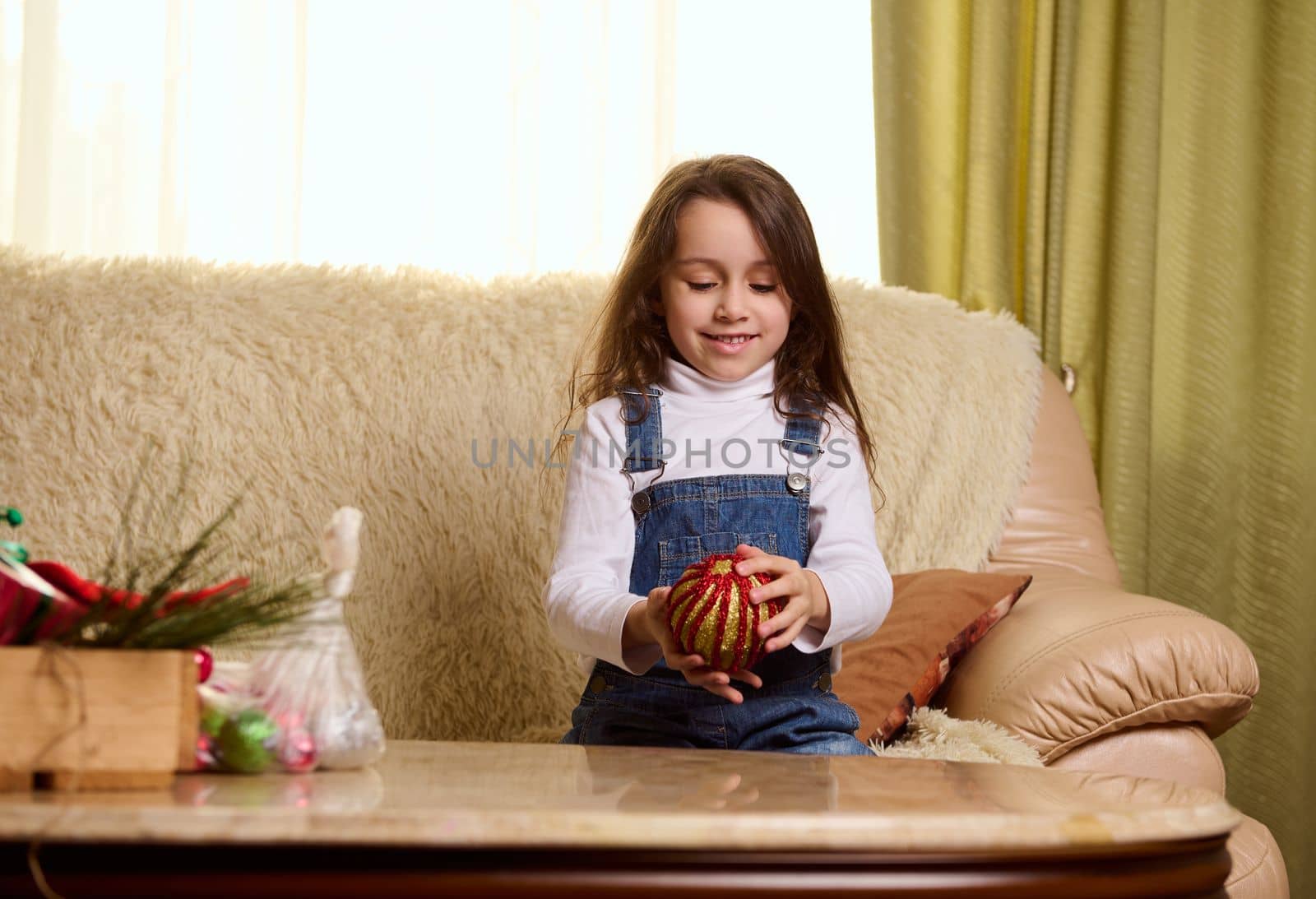 Adorable Caucasian little child girl in blue denim overalls, sitting on a comfortable sofa in the living room, surrounded by festive decorations for upcoming winter holidays and Christmas. Magic mood