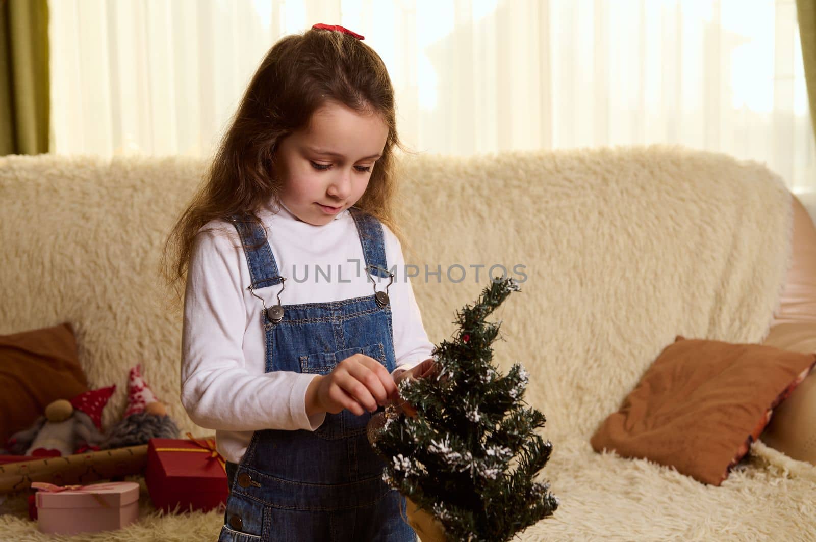 Beautiful Caucasian little child girl decorating a small toy Christmas tree in the home interior. Scandinavian gnomes and gifts on the sofa on the background. Xmas. New Years preparations. December 25