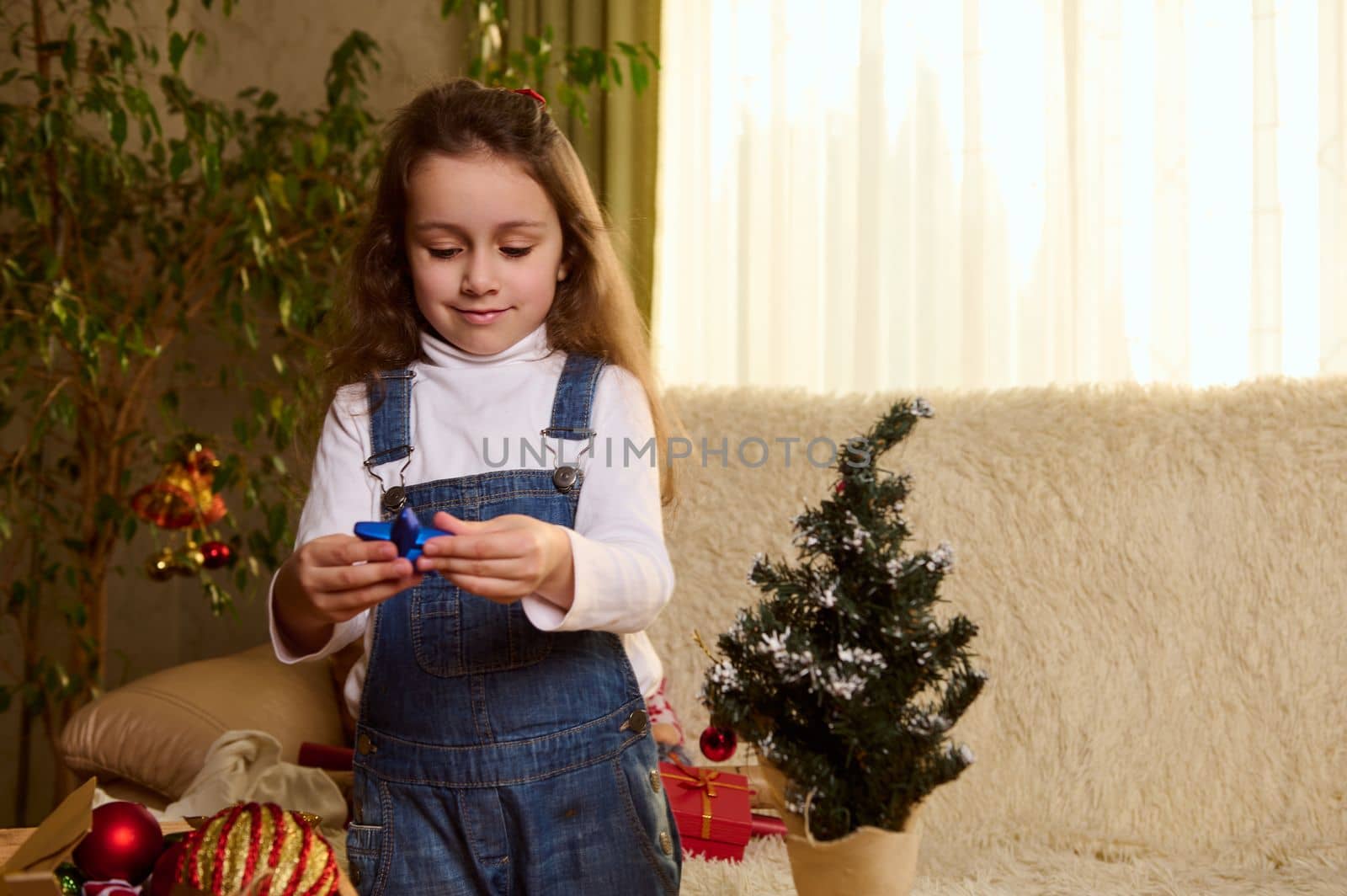 Close-up portrait of charming Caucasian little child girl holding Christmas toys, enjoying New Year and Christmas preparations in the home interior. Happy atmosphere of winter holidays. December 25th