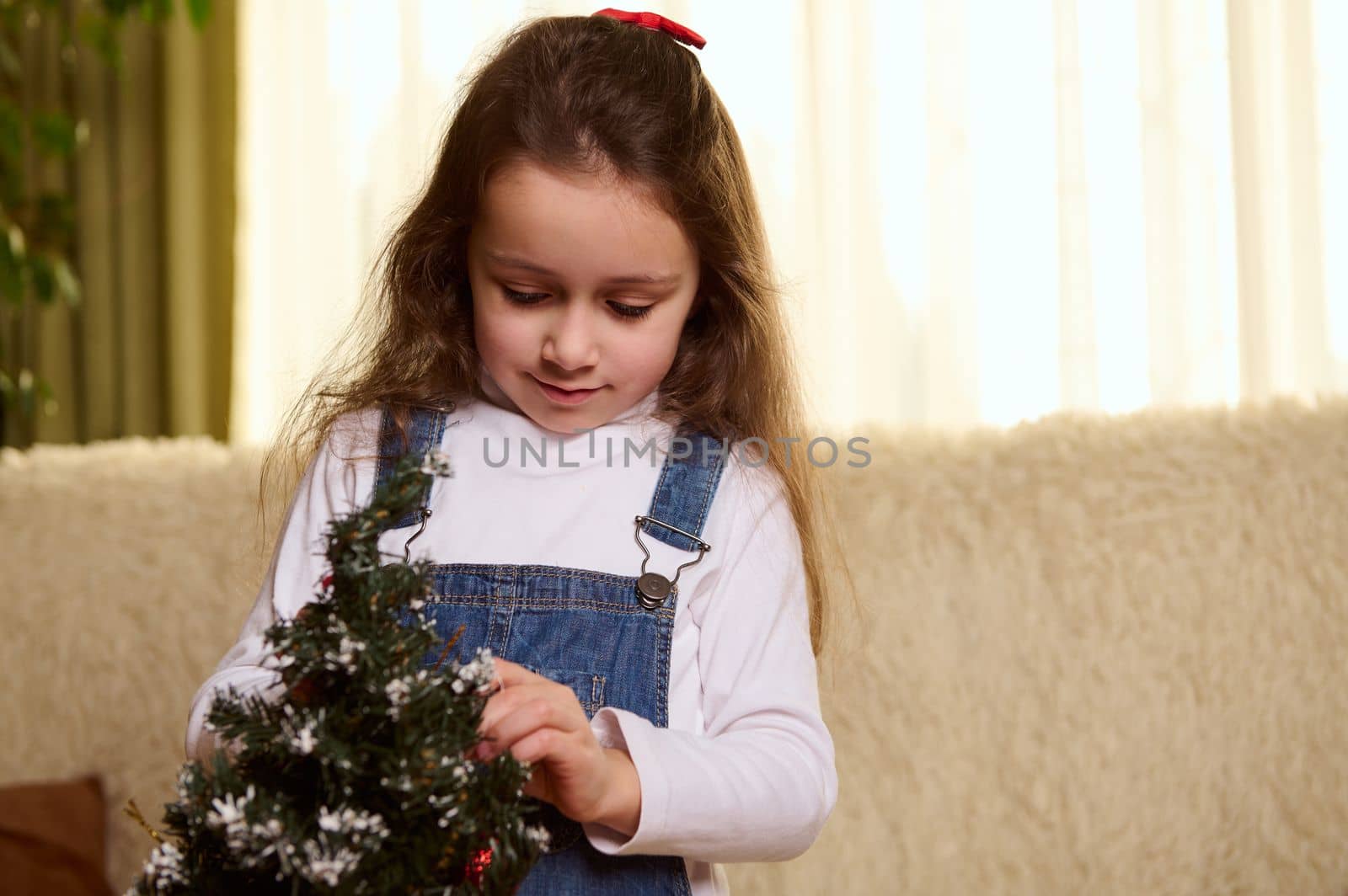 Close-up portrait of Caucasian pretty kid, beautiful little girl with long hair, enjoying decorating a Christmas tree with toys in the home interior. Christmastime. New Year preparations. Festive mood