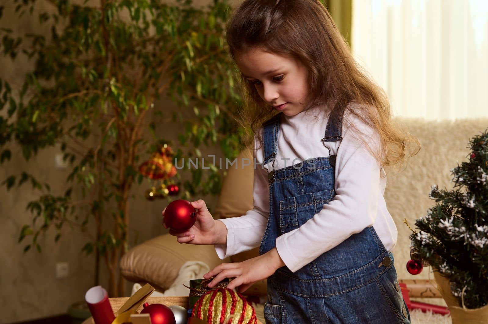 Charming Caucasian little girl with Christmas toys, enjoying the upcoming winter holidays, decorating a toy Xmas tree, in a cozy home interior. Atmospheric mood. Christian traditions. December 25.