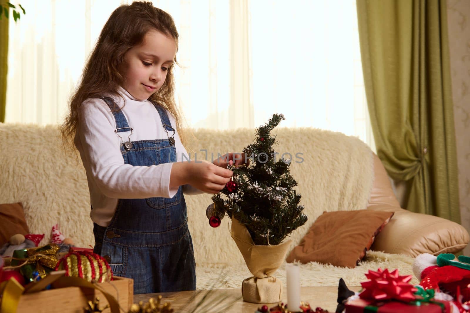 Beautiful Caucasian child, a little girl hanging shiny balls and toys on a small Christmas tree, while decorating home for winter holidays. Merry Xmas and happy New Year. Magic festive atmosphere