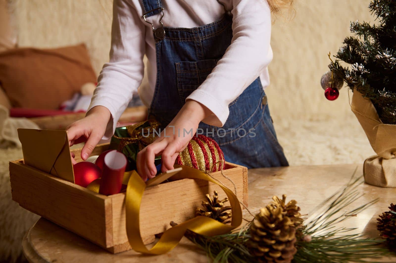 Cropped view of a cute child choosing Christmas toys in a wooden crate for decorating Xmas tree at home. A branch of fir-tree and golden pine cones as a festive ornament, on the table on foreground