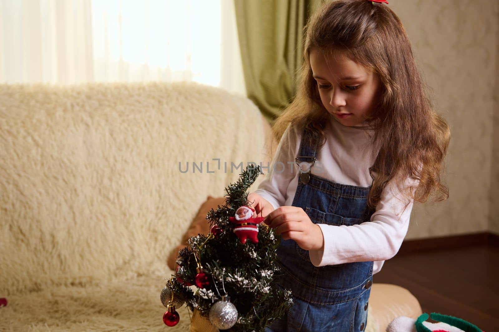 Adorable little girl holding shiny ball while hanging decorative Xmas toys on a Christmas tree in a cozy home interior. by artgf
