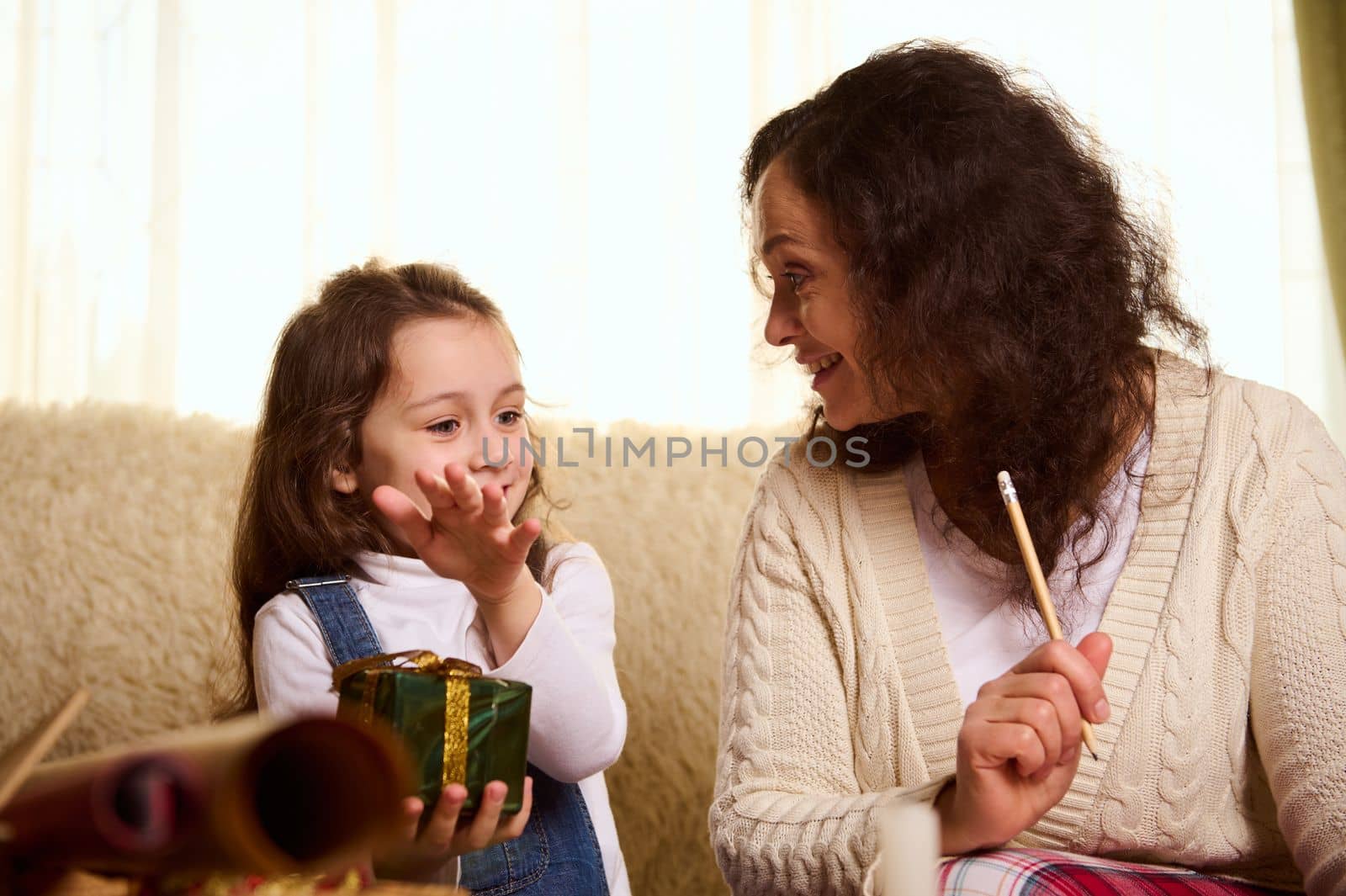 Happy multi-ethnic family of a loving mother and her beloved little daughter writing a gift list, wrapping beautiful presents for the upcoming winter holidays. Merry Christmas. New Year preparations