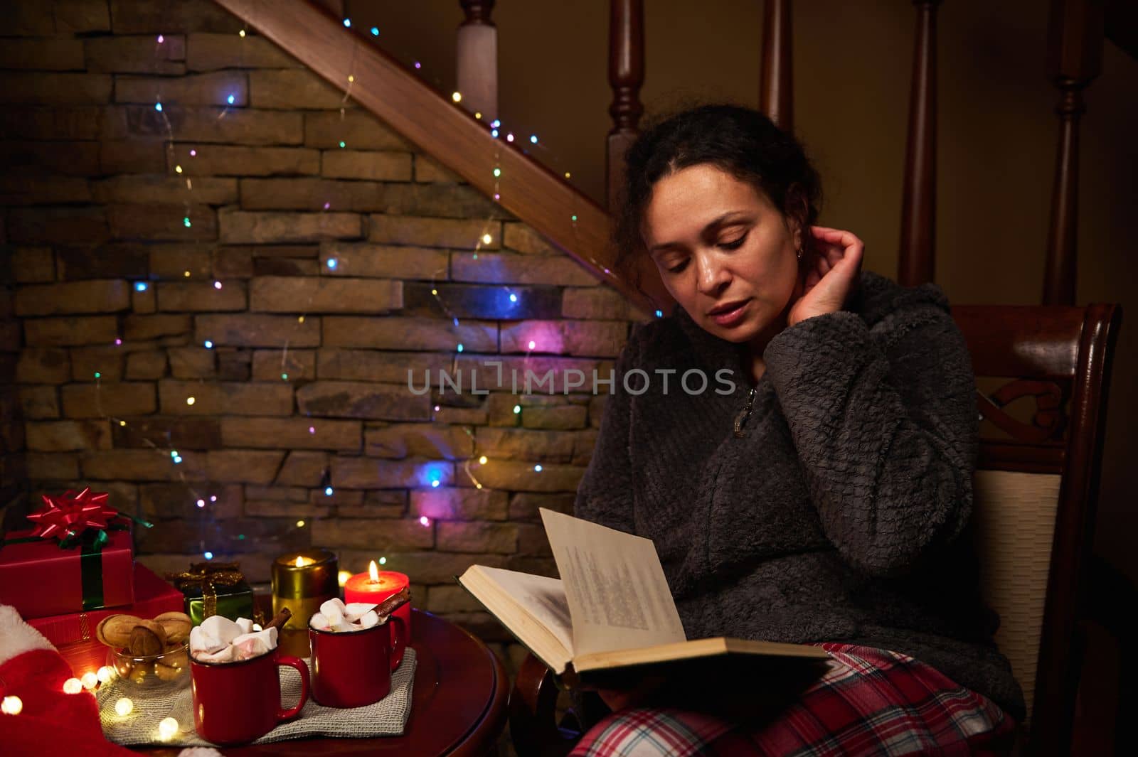Relaxed woman reads book in candlelight, sitting by table with cups of hot cocoa drink with marshmallows, Christmas gifts, beautiful Xmas lights on Santa's hat and sparkling garlands on a brick wall.