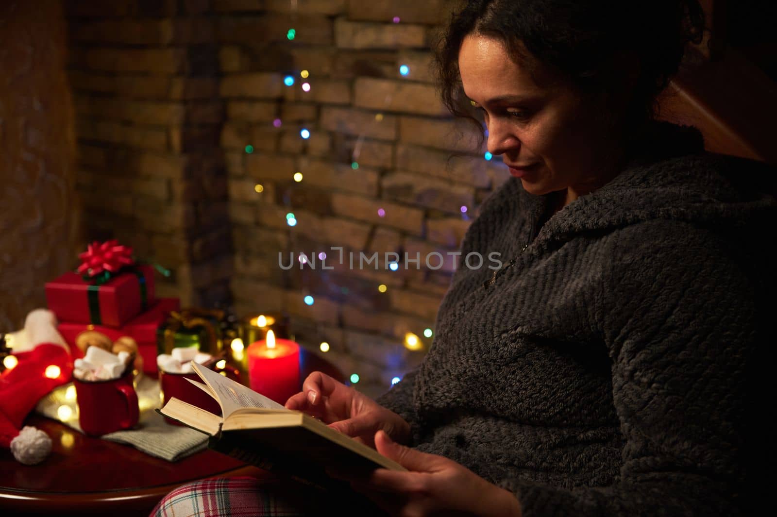 Close-up: Relaxed pretty woman reading book by candlelight, sitting at a festive table with Xmas presents, cups of hot cocoa with marshmallows and sparkling garlands. Merry Christmas. Winter holidays