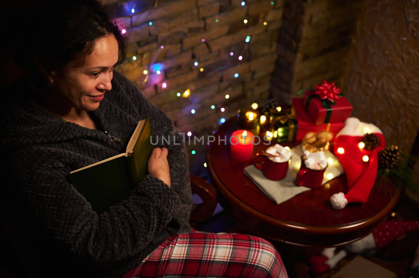 Top view of delightful charming woman with book, enjoying reading by candlelight, sitting at table with Christmas gifts, cups of hot cocoa with marshmallows and sparkling garlands. Merry Christmas.