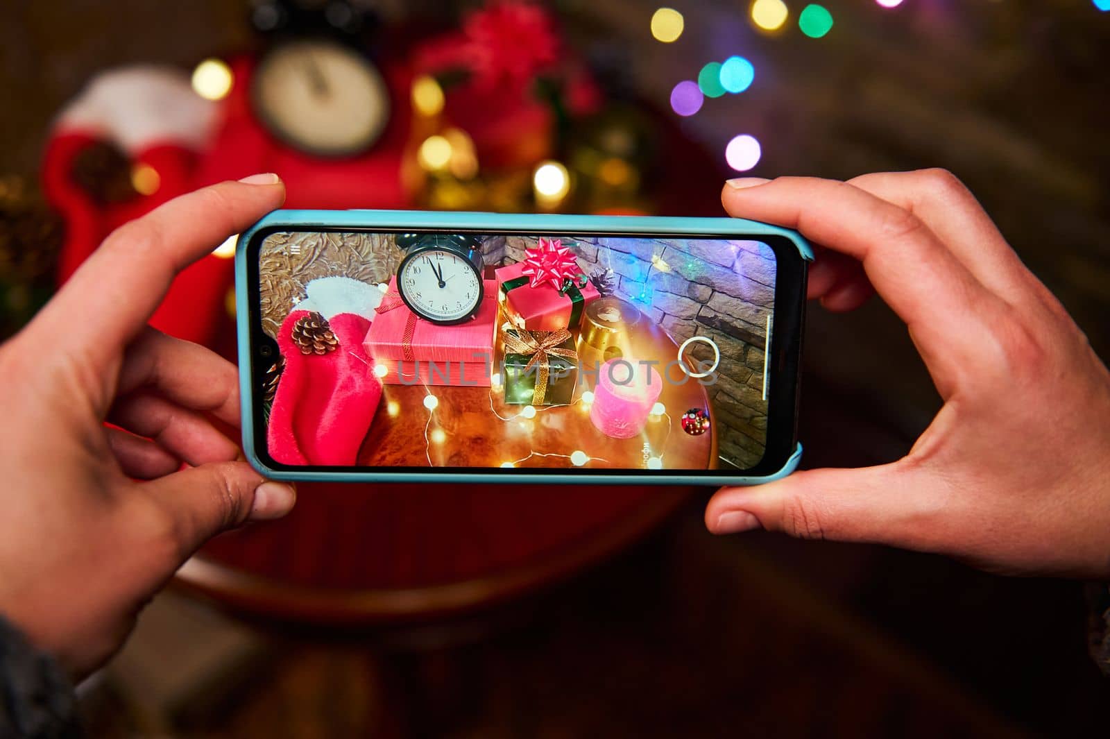 Mobile phone in live view. Female hands hold a smartphone, photograph a cozy Christmas interior with a black alarm clock with midnight on the dial, luminous garlands, lit candles and gifts on table