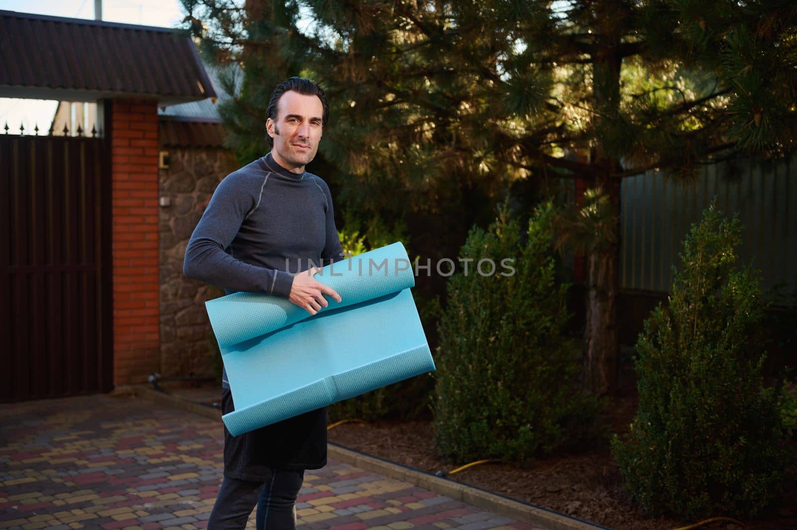 Handsome active Caucasian male athlete, sportsman, fit guy in his 40s holding a fitness mat, working out outdoors at sunset, smiling looking at camera, standing in the house backyard