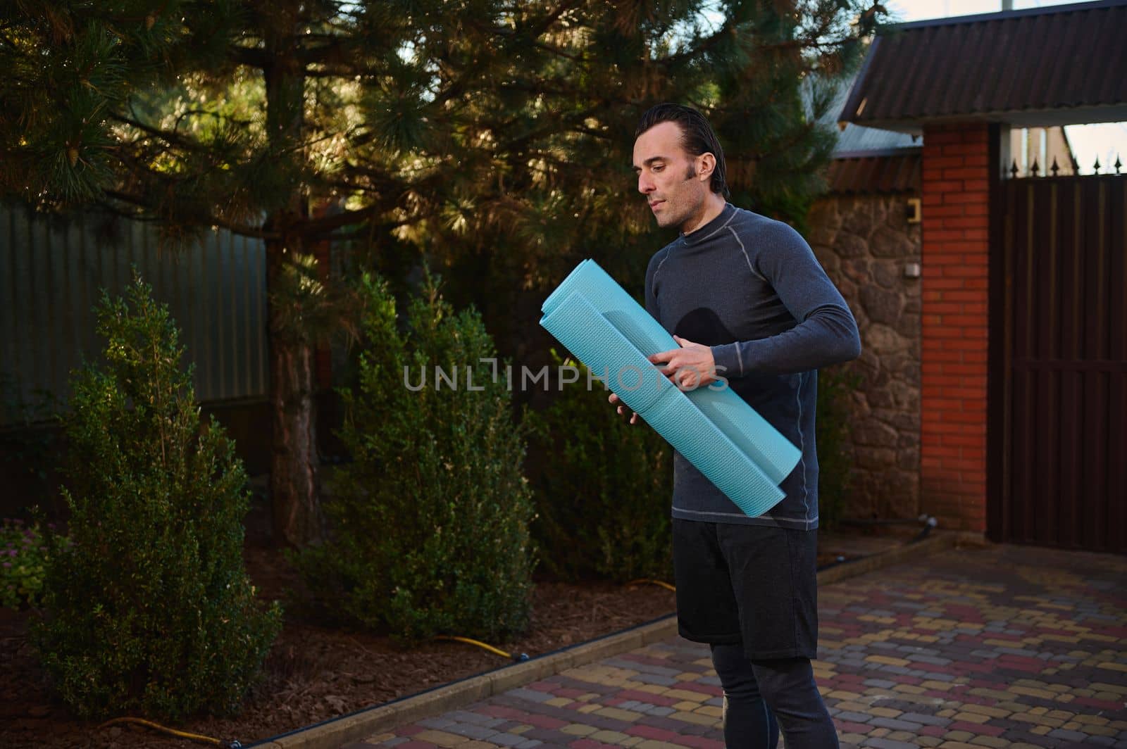 Portrait of a Caucasian man, a determined athlete, sportsman with a fitness mat while exercising outdoors, practicing yoga at sunset. Sport, active and healthy lifestyle.