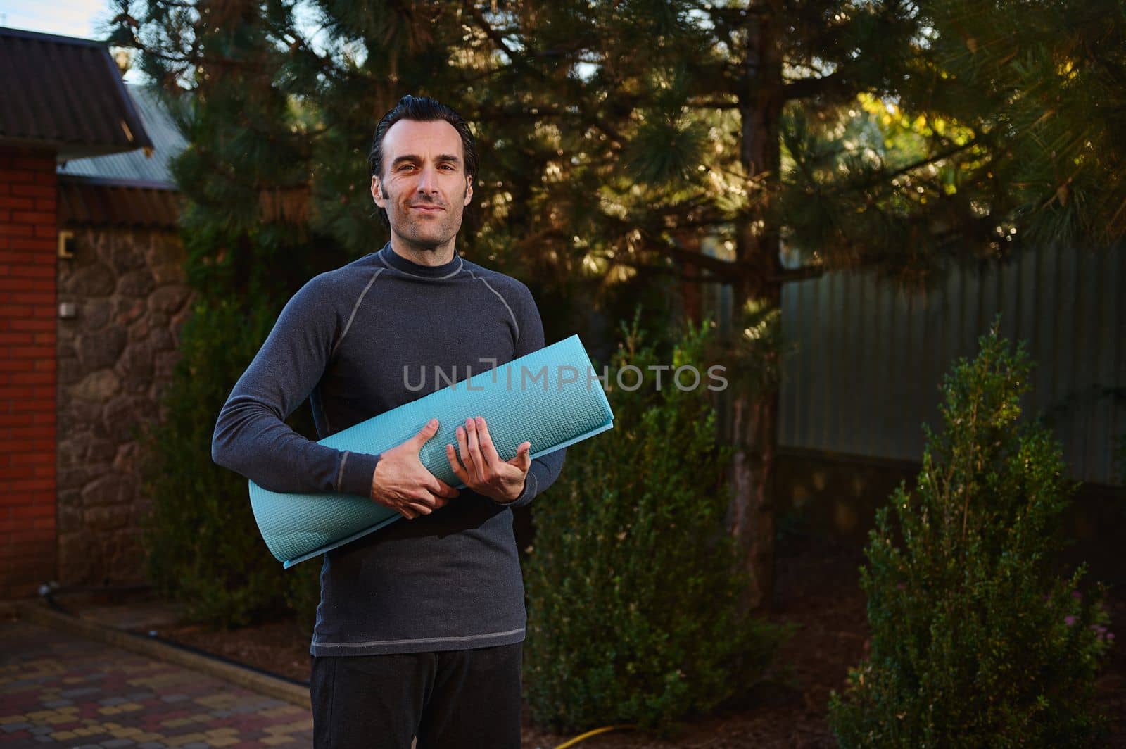 Middle-aged Caucasian handsome active man in sportswear, posing with a blue fitness mat on the backyard, while practicing yoga at sunset. Determined male athlete smiles looking at the camera