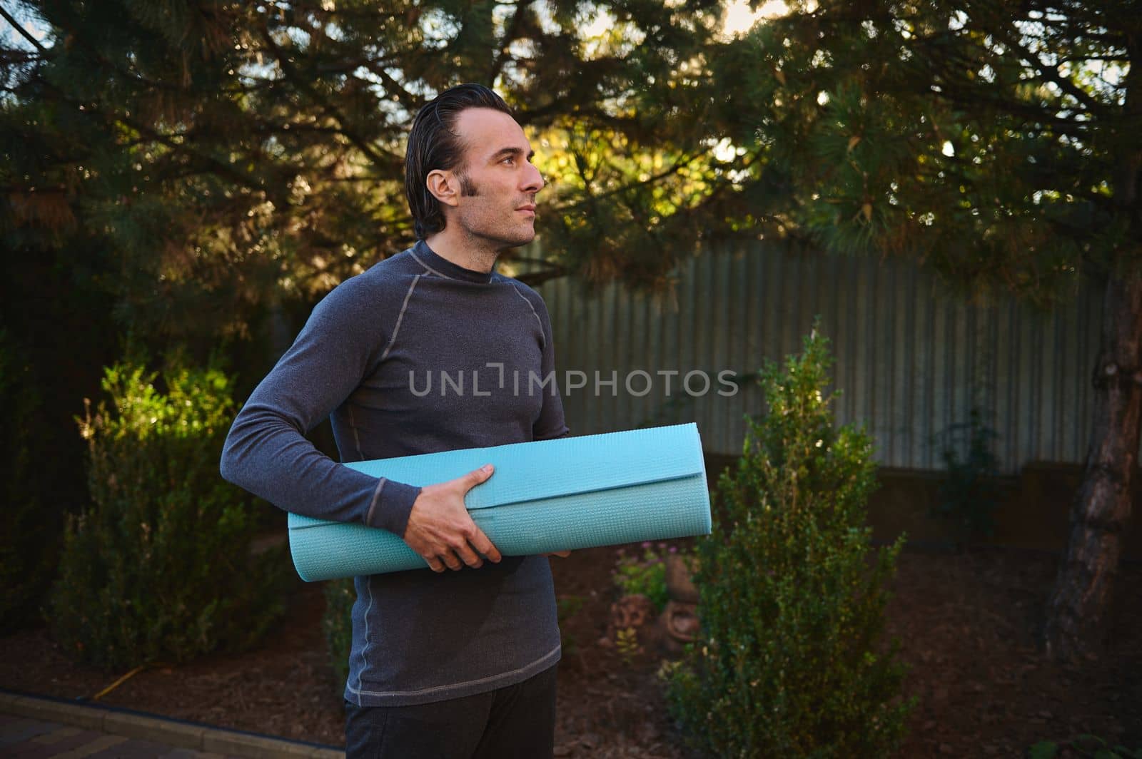Side portrait of a Caucasian handsome active man in sportswear, posing with a blue fitness mat on the backyard, while practicing yoga at sunset. Attractive muscular male athlete working out. Sport