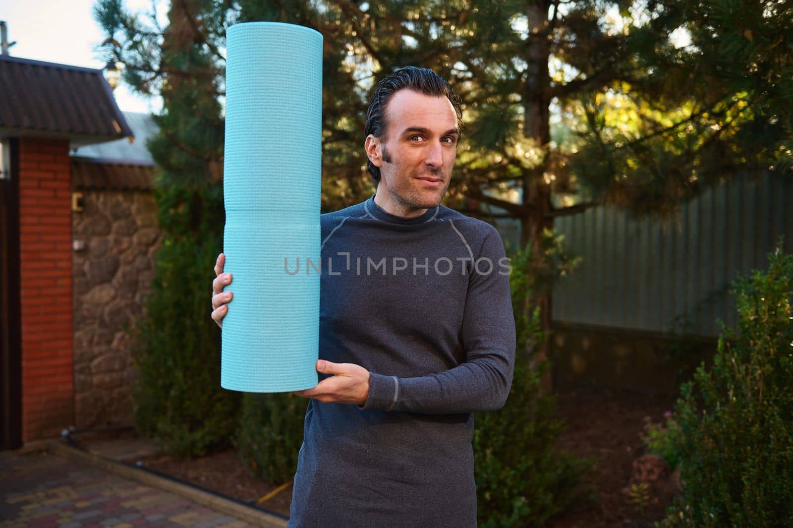 Portrait of an active Caucasian man, a determined athlete, sportsman with a fitness mat, smiles looking at the camera while exercising outdoors, practicing yoga at sunset. Sport and healthy lifestyle.