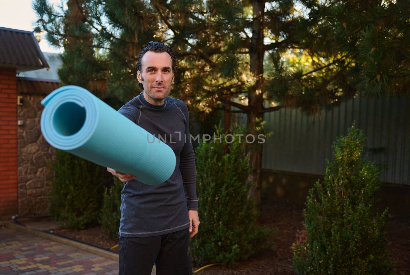 Attractive young Caucasian man, determined athlete, sportsman, yogi in gray activewear, looks at camera while handing a rolled blue fitness mat. Yoga. Sport concept. Active lifestyle. Body conscious