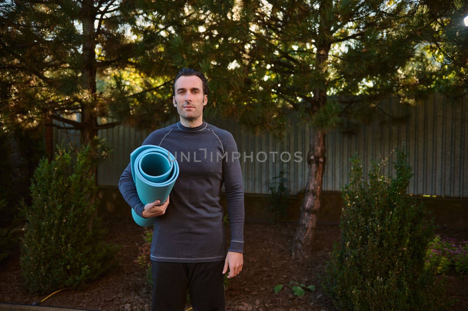 Handsome active man athlete, sportsman with a fitness mat looking at the camera while practicing yoga outdoors at sunset by artgf