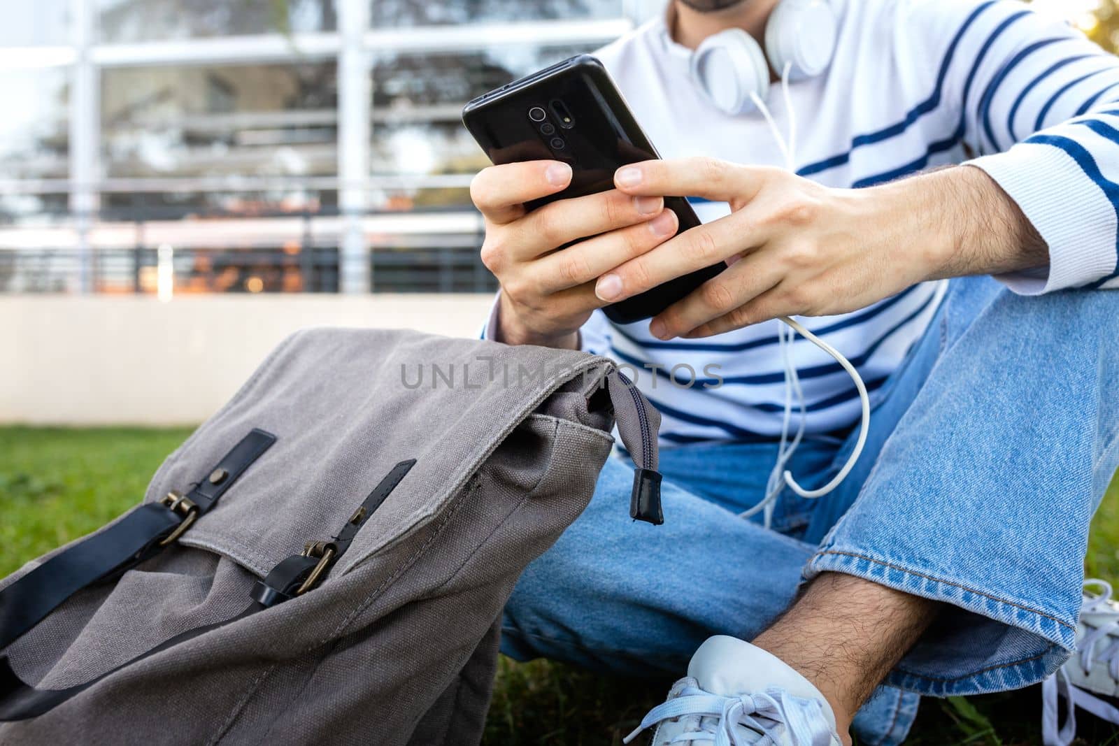 Close up of unrecognizable male hands using mobile phone outdoors. College student sitting on grass in campus texting message with phone. Education concept.