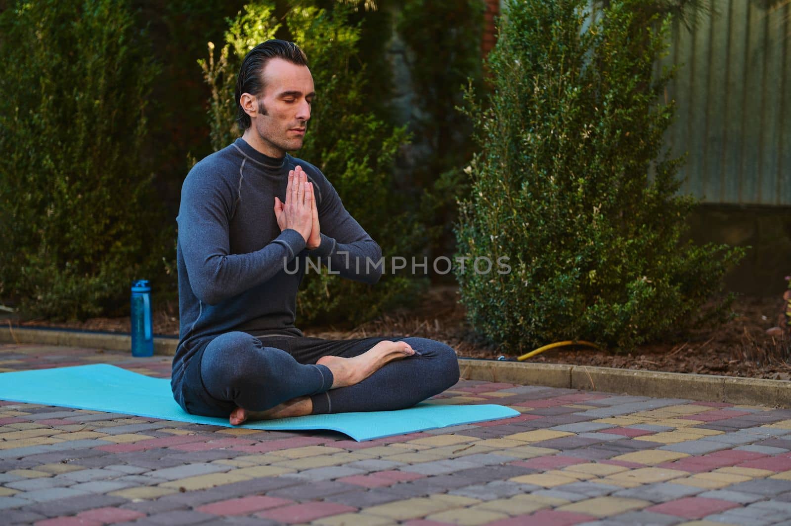 Handsome peaceful and confident man athlete, yogi, sitting barefoot in lotus pose on a fitness mat, keeping hands palms together, meditating during yoga practice at sunset, outdoors. Spiritual growth