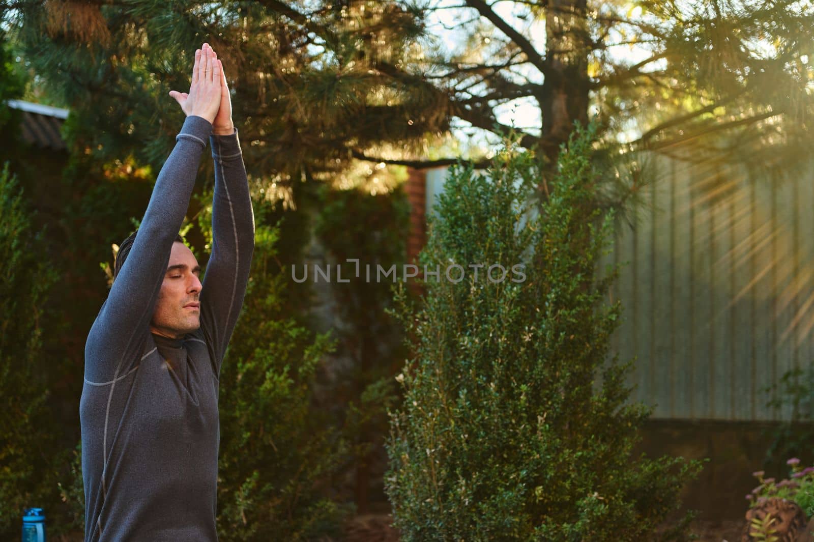 Active middle-aged Caucasian sportsman, male athlete, yogi raising hands to the sky, palm to palm, meditating, stretching his body, doing yoga at sunset, outdoors against coniferous trees background