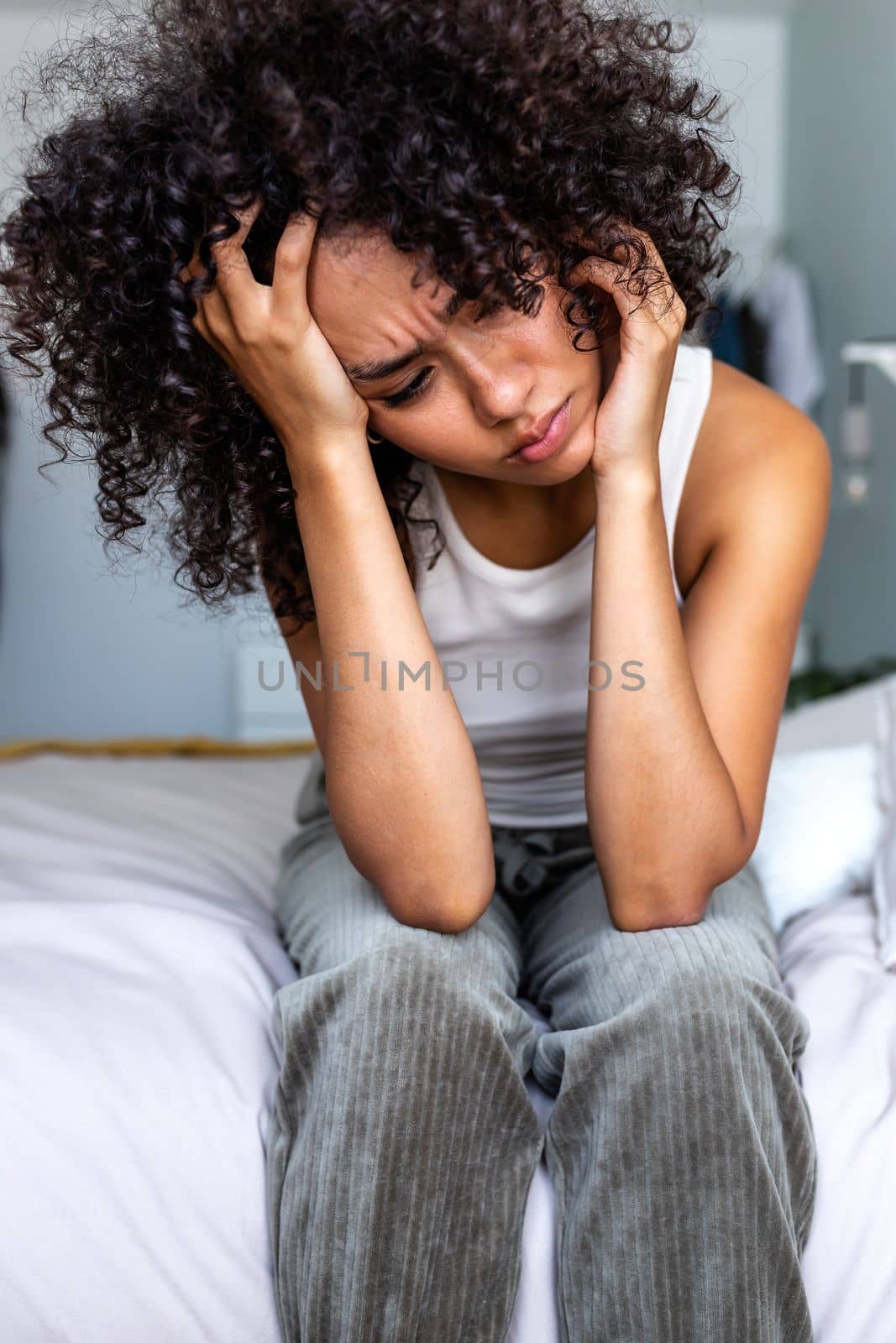 Vertical portrait of young african american latina woman sitting on bed ledge feeling depressed and sad. by Hoverstock