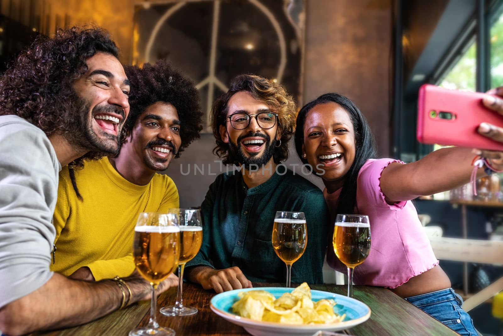 African American young woman taking selfie with group of friends using phone in a bar enjoying some beer and food. Lifestyle concept.