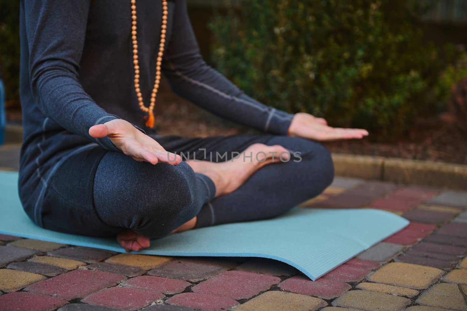 Cropped view of male athlete, yogi wearing rosary beads and gray sportswear, sitting on fitness mat in lotus position and palms up on knees, practicing yoga. Prayer, gratitude. Yoga pose. Meditation.