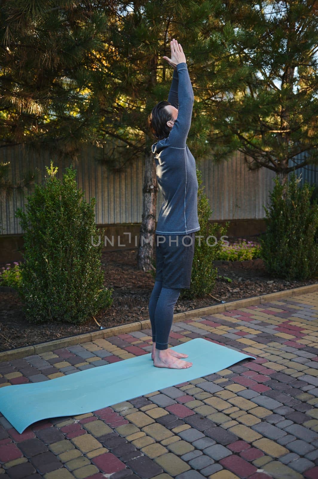 Full-length portrait of a sportsman doing outdoor sports, standing barefoot on a fitness mat, stretching his body, pulling his arms up during stretching and yoga practice. Active healthy lifestyle.